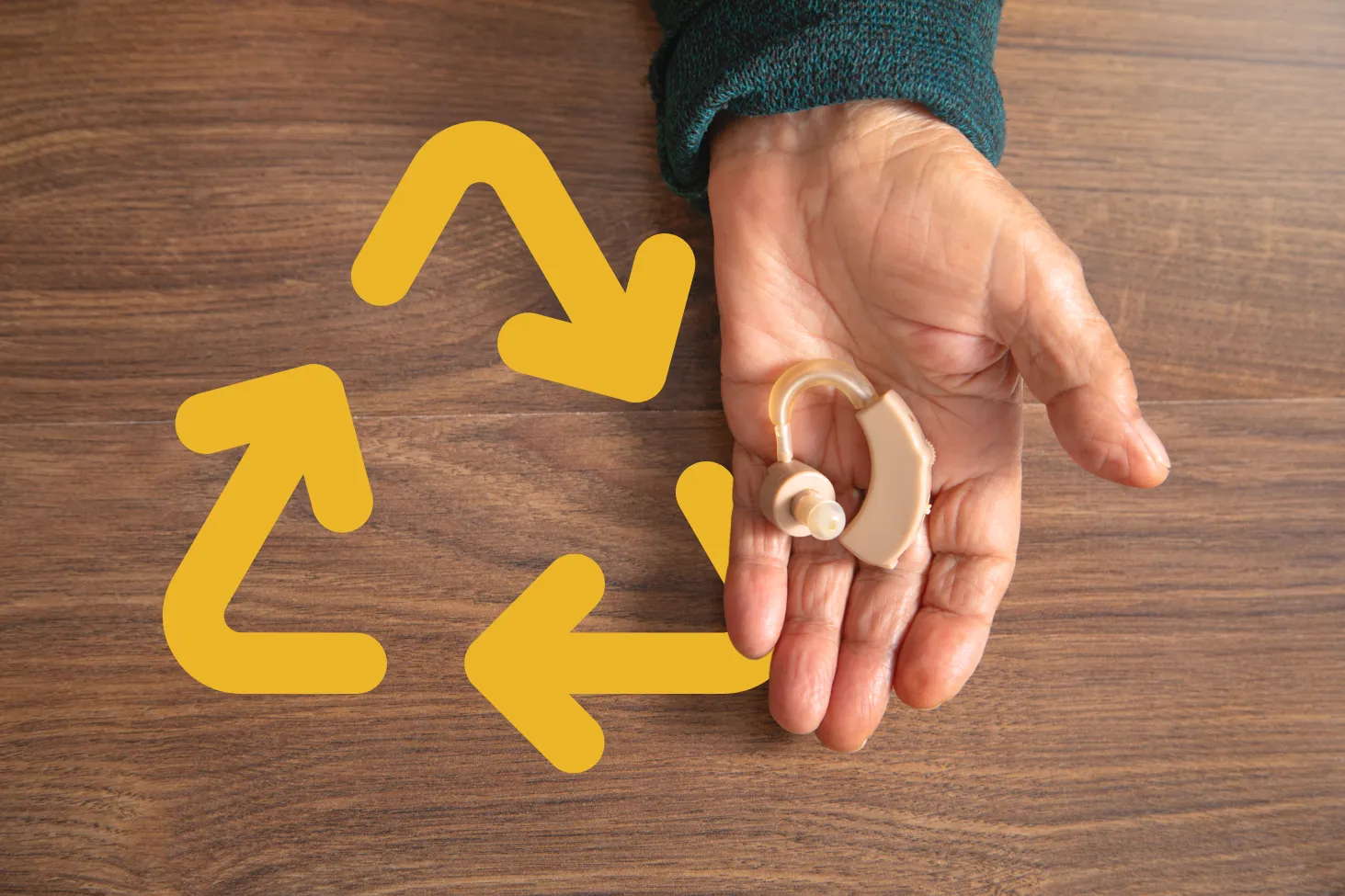 Hand holding a hearing aid above a wooden table with a yellow recycling symbol